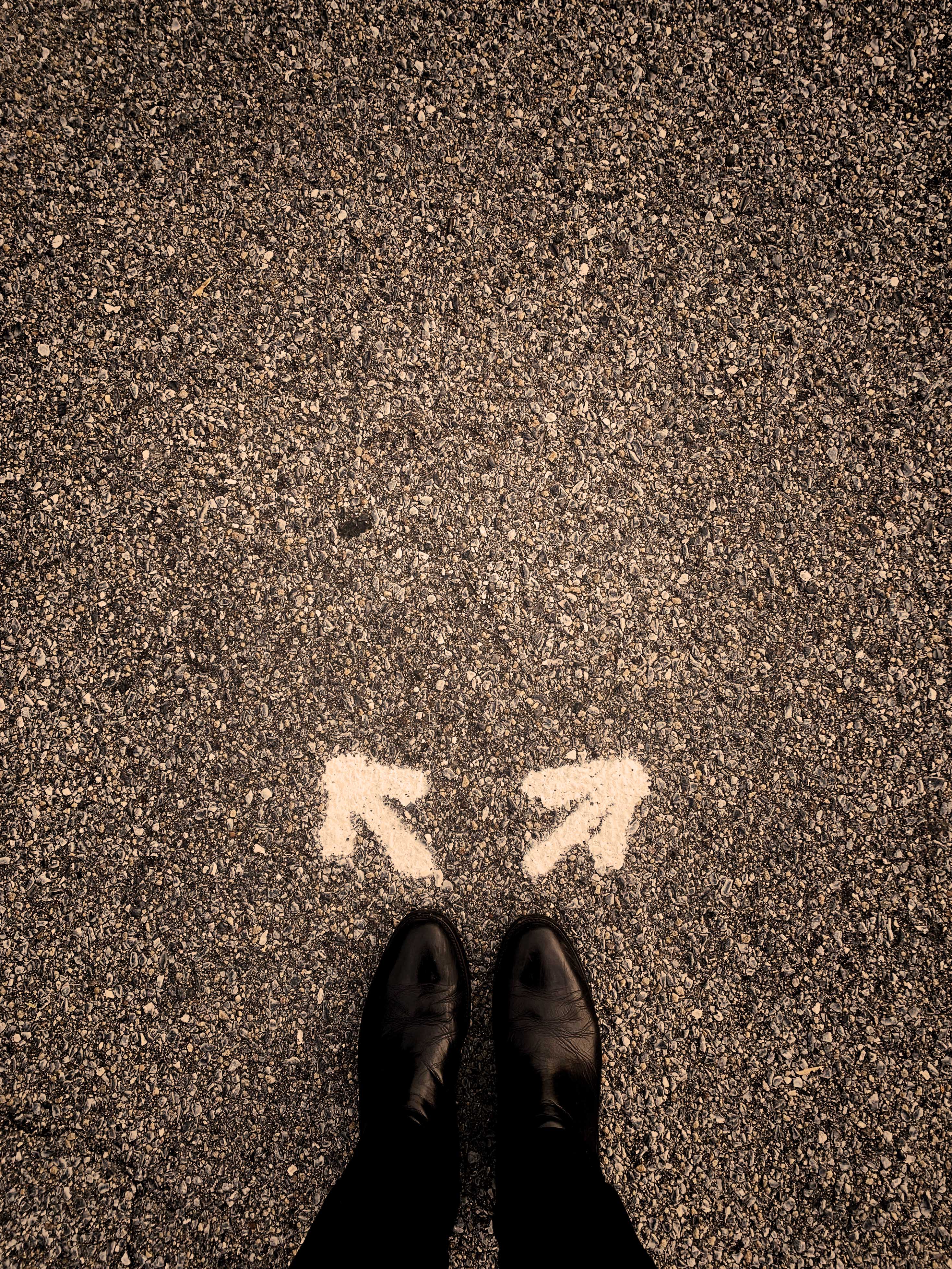 Image to indicate the concept of 'choice'. Sepia toned image of a person standing on gravel wearing Oxford shoes with both feet pointing straight ahead. A pair of arrows are drawn, one in front of each foot, pointing in opposing directions.